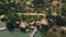 Exotic bungalows with thatched roof and boat pier on shore green lake aerial view. Drone view holiday cottages on shore