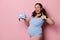 Expectant mother holds newborn clothes on her hand, winks and smiles a toothy smile at camera, isolated pink background