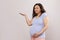 Expectant mother stroking her belly, holding copy space on her palm up, looking at camera, isolated on white background