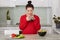 Expectant young mother has breakfast at kitchen, eats fresh vegetable salad and drinks tea, wears casual red sweater, has healthy