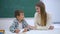 Experienced tutor helps to learn to schoolboy at table near board in classroom of School