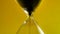 Extreme close-up of black sand running down through hourglass on a yellow background. A concept the sand runs out to the