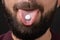 Extreme close-up of white round pill on tongue of bearded man face with dark mustache on black background, take vitamins