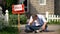 Extremely depressed man sitting on ground by sold signboard, financial problems