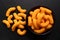 Extruded cheese puffs in a black ceramic bowl next to spilled cheese puffs isolated on black. Top view