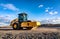 Eye catching yellow road roller with enclosed climate controlled cabin stands on not ready new road, stones, blue sky, clouds,