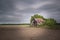 Eye-level shot of old wooden decrepit barn near trees and bushes under dark, cloudy sky