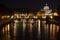 A fabulous view of night Rome with a church, a bridge, a ship and the light of lanterns reflected in the Tiber.