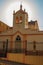 Facade of small church and belfry, behind iron fence, with sunshine behind at sunset in SÃ£o Manuel.