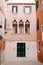 Facade of a three-story peach house in Venice, Italy. Massive green wooden front door, three classic Venetian windows