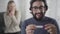 Face of shocked Caucasian man holding pregnancy test, looking back at his wife or girlfriend clapping hands, and smiling