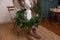 Faceless young woman sits on chair, holding in hands asymmetrical Christmas wreath. Scandi design