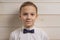 A fair-haired boy of 10 years in a white shirt with the self-tie bow tie smiles against the background of a wooden wall