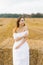 A fair-haired young woman in a white dress in a field with stacks of straw is enjoying a summer day