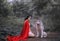 Fairy tale about red cap, dark-haired girl on ground in thick forest in short white light dress, long scarlet cloak