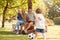 Family With Baby Girl Having Fun In Park Playing Football And Sitting On Seat Under Tree