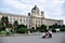 A family, with a child sitting in the seat and other people, stroll in front of the Kunst Museum in Vienna. Vienna, the capital of