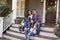 Family With Children Sit On Steps Leading Up To Porch Of Home