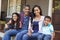 Family With Children Sit On Steps Leading Up To Porch Of Home