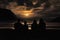 Family of four sitting on ocean shore and looking at dramatic orange sunset at dusk, rear view