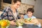 Family at home standing in kitchen together son putting cucumbers into salad joyful helping mom