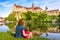 Family sit near medieval Sigmaringen Castle, Germany. Young woman and her baby are by river in summer