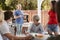 Family at a table outdoors turn to dad standing by barbecue