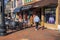 A family walking along a red brick sidewalk lined with shops, restaurants and bars with tables and chairs on the sidewalk, parked