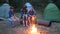 Family weekend in forest, mom and kids communicate near bonfire, young family in background tents and trees,