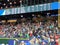 Fans in bleachers do the wave with scoreboard above