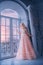 Fantasy woman princess stands near window castle white room. Girl looking outdoor with hope