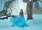 Fantasy young woman in image of medieval princess sits on a tree in luxurious blue vintage dress. Background Winter