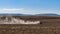 Far view of tractor plows hilly field raising a large cloud of dust after a dry summer season and blue sky in background