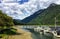 A faraway view from the shore of a beautiful marina full of boats and old inn, surrounded by forest and mountains