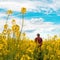 Farm worker wearing red plaid shirt and trucker`s hat standing in cultivated rapeseed field in bloom and looking over crops