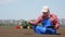 Farmer, agronomist sits between soil rows, checking quality of soil on farm field. background of working tractor