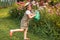 Farmer girl in summer straw hat. Little gardener farming, watering flowerbed with pink flowers, having fun in garden. Big green