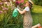 Farmer girl in summer straw hat. Little gardener farming, watering flowerbed with pink flowers, having fun in garden. Big green