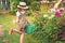 Farmer girl in summer straw hat. Little gardener farming, watering flowerbed with pink flowers, having fun in garden. Big green