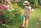 Farmer girl in summer straw hat. Little gardener farming, watering flowerbed with pink flowers, having fun in garden. Big green