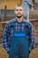 Farmer handsome european appearance male rural portrait with beard, shirt and overalls with hands in pockets looking at camera
