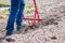 A farmer in jeans digs the ground with a red fork-shaped shovel. A miracle shovel, a handy tool. Manual cultivator. The cultivator