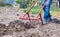 A farmer in jeans digs the ground with a red fork-shaped shovel. A miracle shovel, a handy tool. Manual cultivator. The cultivator