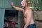 Farmer leaning on a industrial shed. Man is bald with a beard and no shirt