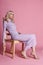 Fashion Beautiful blonde woman in knitted closed long dress sitting on a chair on a pink background. Perfect hair and makeup