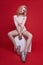 fashion beautiful woman in bodysuit sits on a white cube and poses on a red crimson background. Slender blonde girl