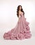 Fashion brunette woman in gorgeous long pink dress posing isolated on white studio background. Beautiful Lady in luxury lush gown.