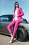 Fashion girl standing next to a retro sport car on the sun. Stylish woman in a pink suit waiting near classic car