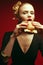 Fashion & Gluttony Concept. Portrait of luxurious red-haired model in black cocktail dress eating burger over red background.