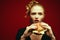Fashion & Gluttony Concept. Portrait of luxurious red-haired model in black cocktail dress eating burger over red background.